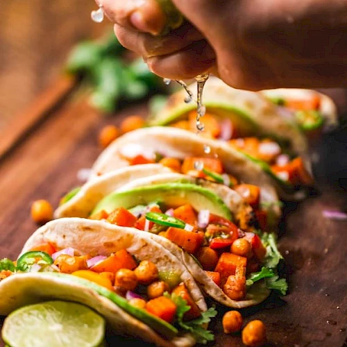 Tacos filled with chickpeas, vegetables, and avocado are lined up on a board. A hand is squeezing lime juice over them.