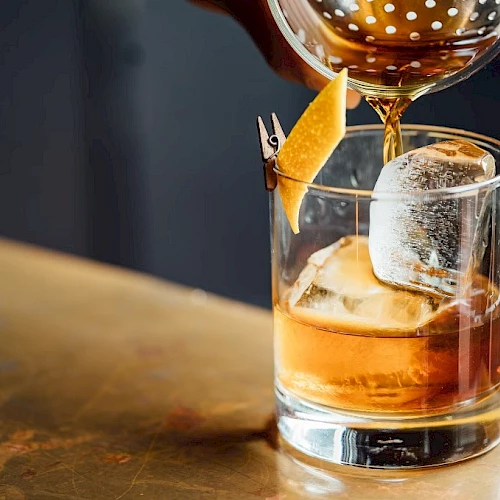 A cocktail is being poured into a glass with ice and an orange peel garnish, on a bar counter.