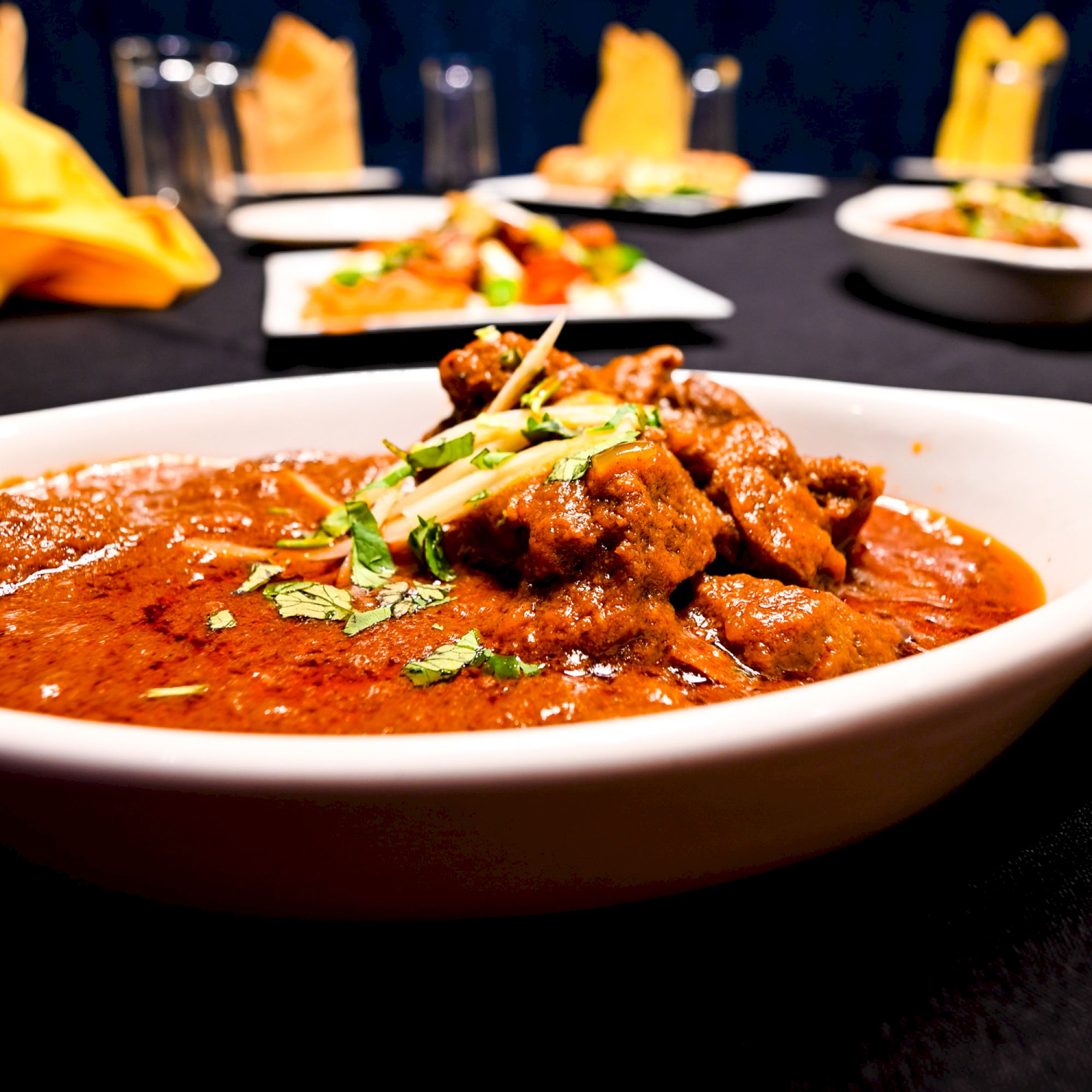 A dish of curry with garnished herbs is served in a white bowl on a table, surrounded by more plates and yellow napkins.