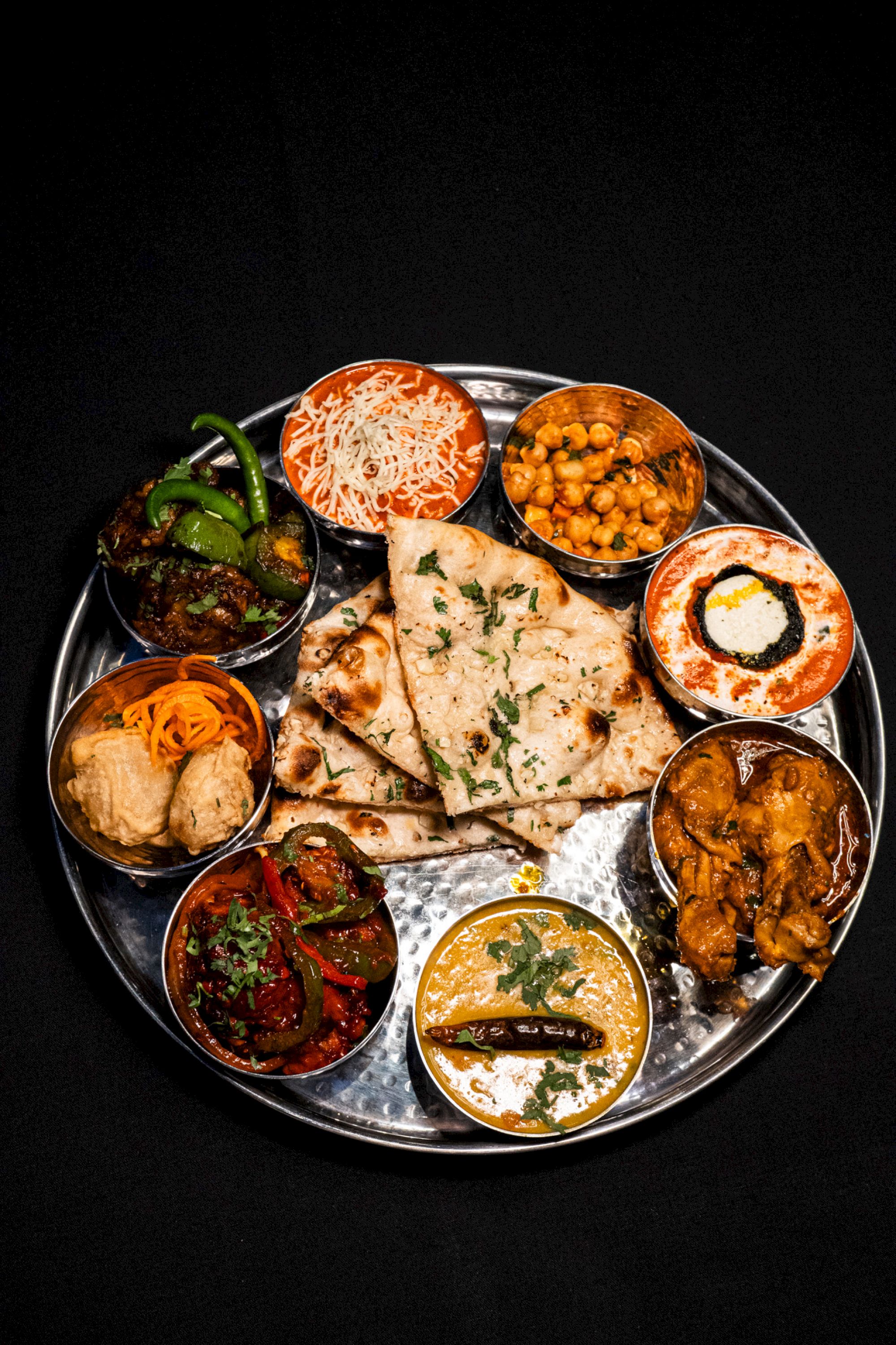 The image shows a platter with naan, various Indian curries, chickpeas, peppers, and more, arranged on a round silver dish.