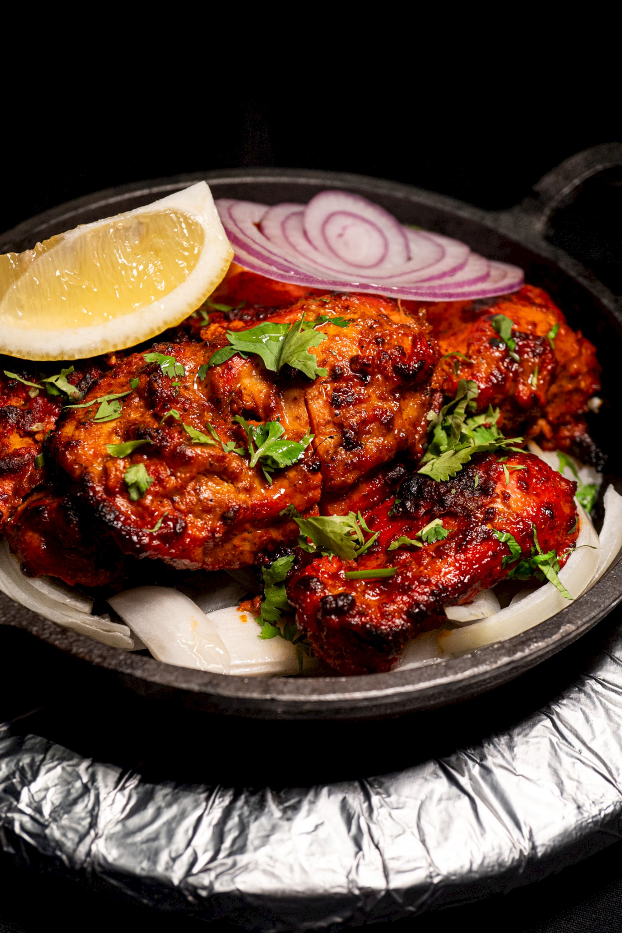Grilled chicken dish with garnished herbs, sliced red onions, and a lemon wedge on top, served in a round skillet.