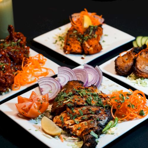 Plates of assorted Indian dishes with grilled meats, garnished with carrots, onions, and spices, are arranged on a dark table.
