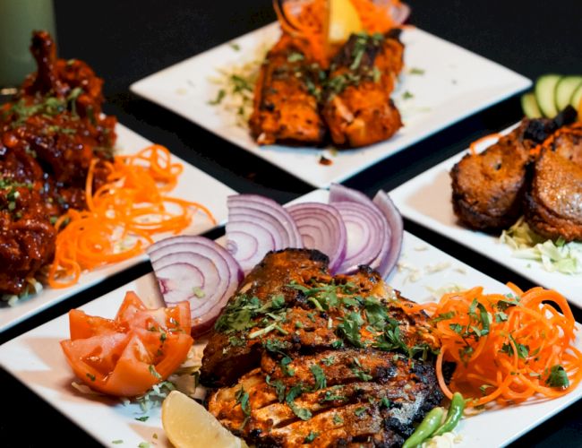 Plates of assorted Indian dishes with grilled meats, garnished with carrots, onions, and spices, are arranged on a dark table.
