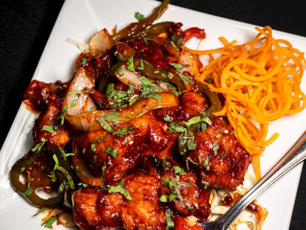 A square plate with saucy chicken, sliced vegetables, garnished with herbs, and spiralized carrots, served with a spoon on the side.