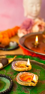 A vibrant festive setup with lit diyas, colorful rangoli, sweets, and a blurred Buddha statue in the background. Ends with a period? Wait. "always ending the sentence." So end with a period. 140 chars max. Let's craft:"Colorful Indian festival scene with glowing diyas, sweets on a decorative rangoli, and a serene Buddha statue blurred in the background."