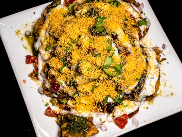 The image shows a plate of Indian chaat with toppings like sev, yogurt, diced tomatoes, and cilantro, served on a white square plate.