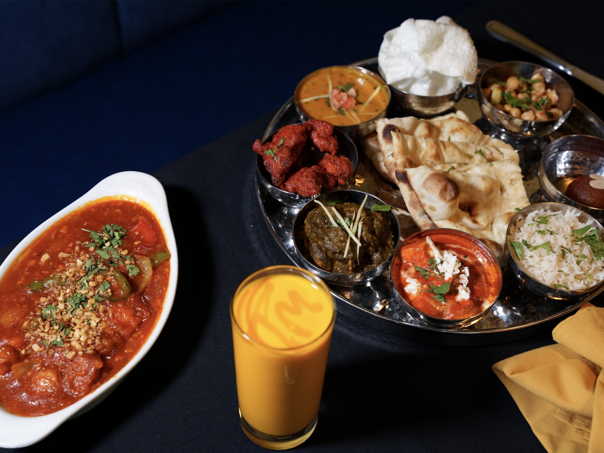 An Indian-style meal platter with naan, rice, mixed curries, fried snacks, chutneys, and a fresh juice drink.