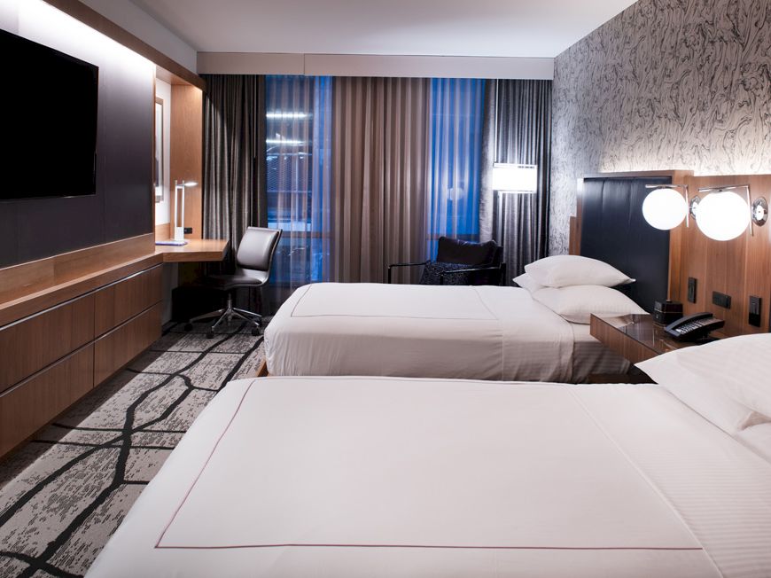 The image shows a modern hotel room with two beds, a large TV, a desk with a chair, and a window with curtains.