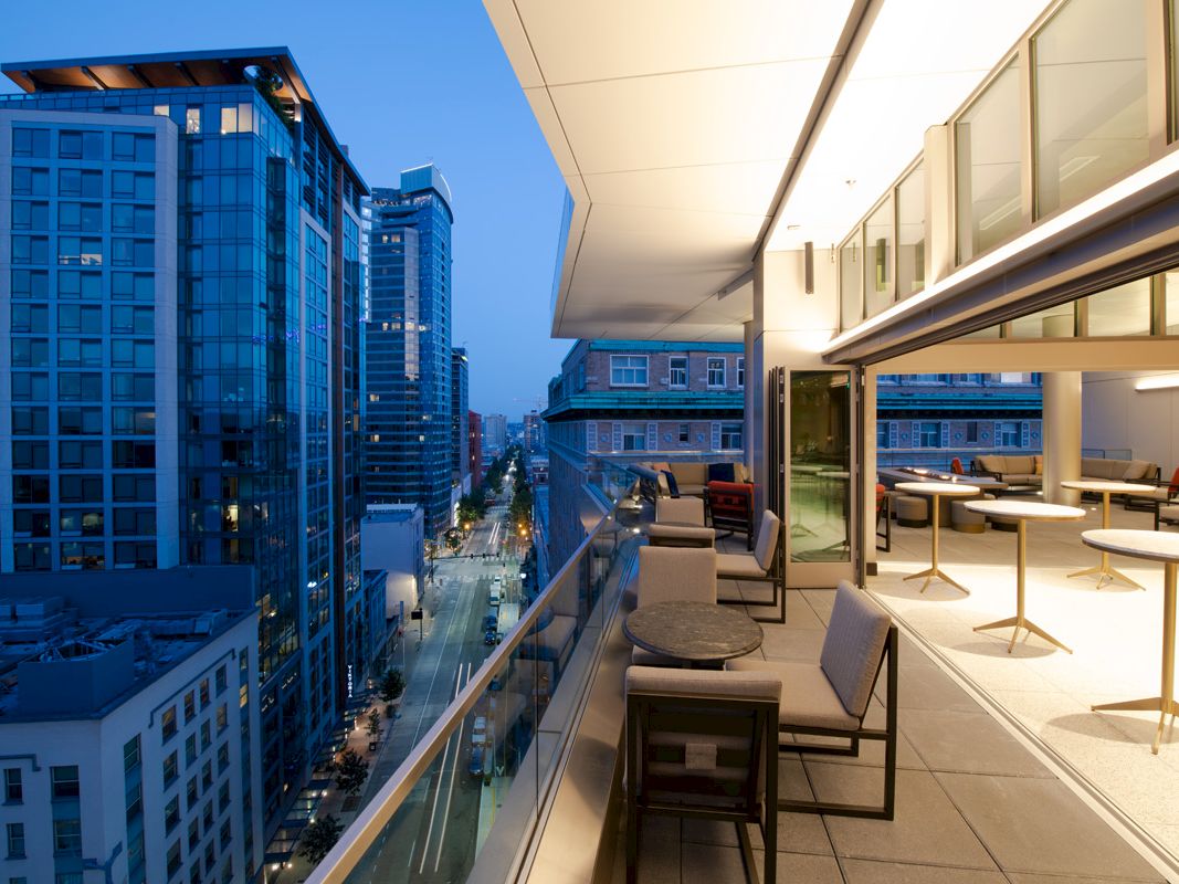A modern rooftop terrace at dusk, featuring seating areas and a cityscape view with tall buildings and a lit-up street.