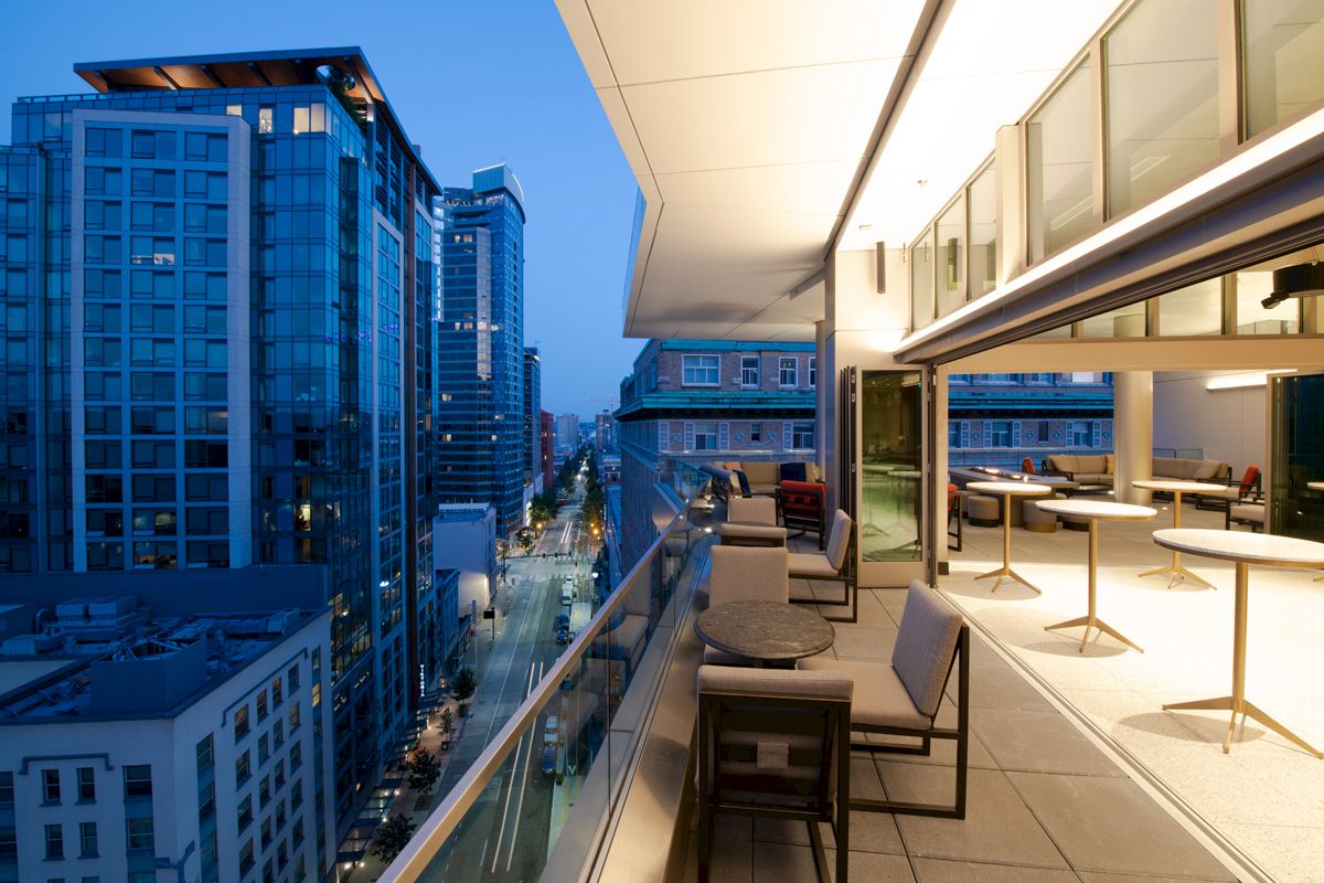 A modern rooftop terrace overlooks a city street lined with tall buildings, featuring outdoor seating and ambient evening lighting.