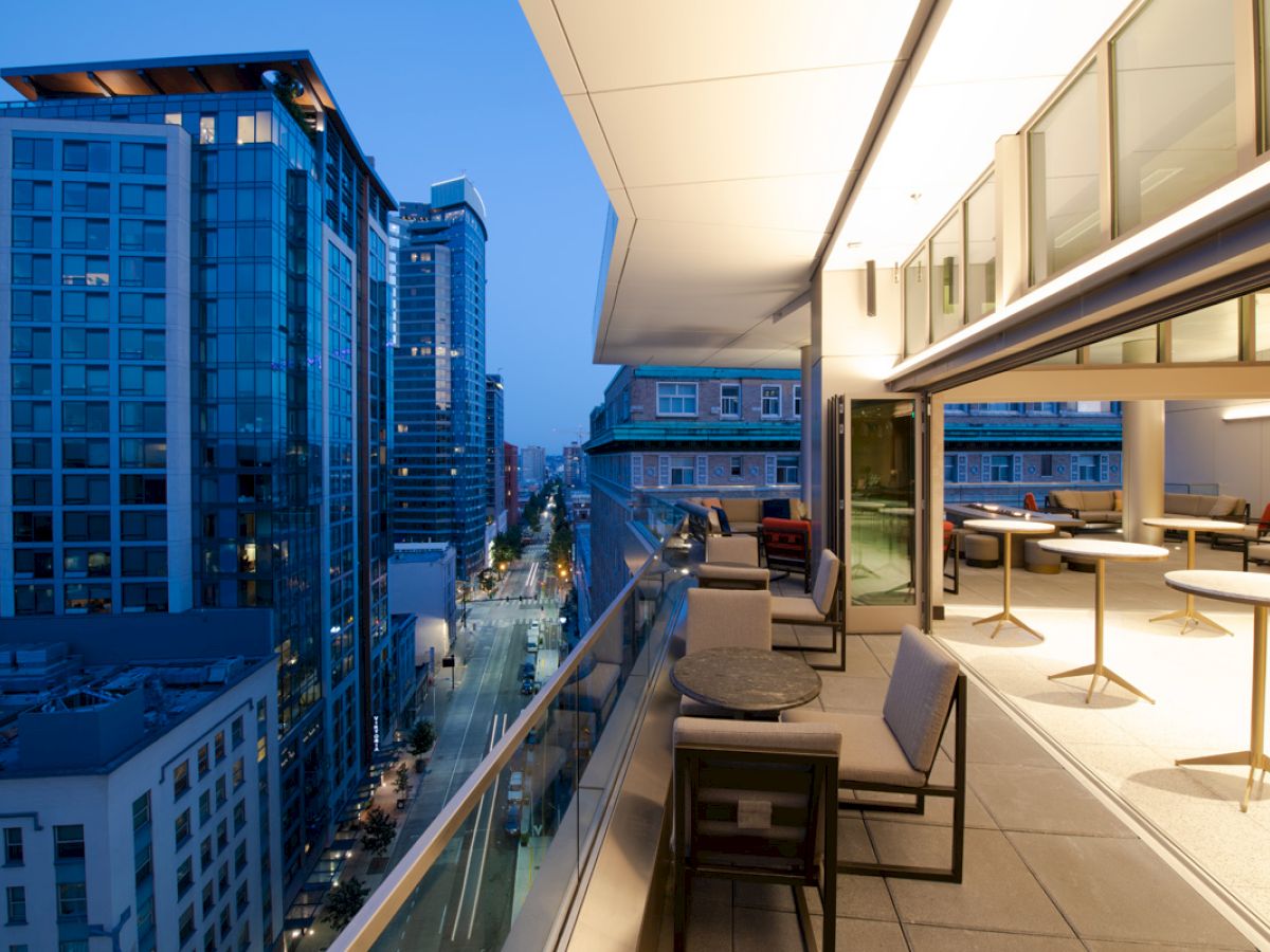 A modern rooftop terrace overlooks a city street lined with tall buildings, featuring outdoor seating and ambient evening lighting.