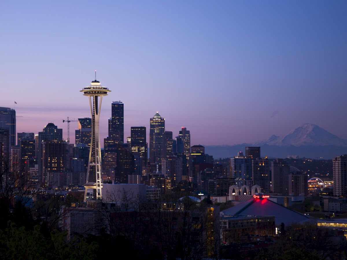 A twilight view of Seattle's skyline, featuring the Space Needle, with a distant mountain visible against a purple-blue sky.