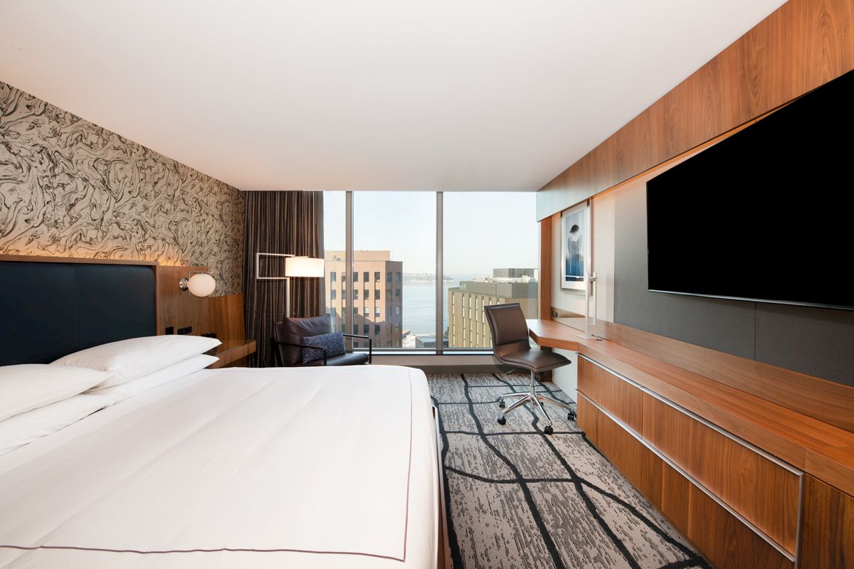 A modern hotel room with a large bed, flat-screen TV, desk, and a window view of buildings.