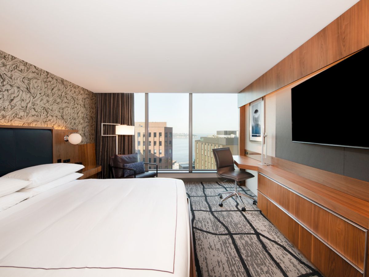 A modern hotel room with a large bed, flat-screen TV, desk, and a window view of buildings.