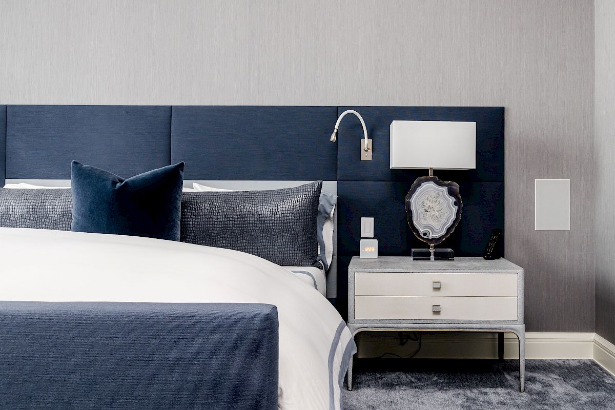 A modern bedroom with a blue and white color scheme, featuring a bed, side table, lamp, and decorative pillows.