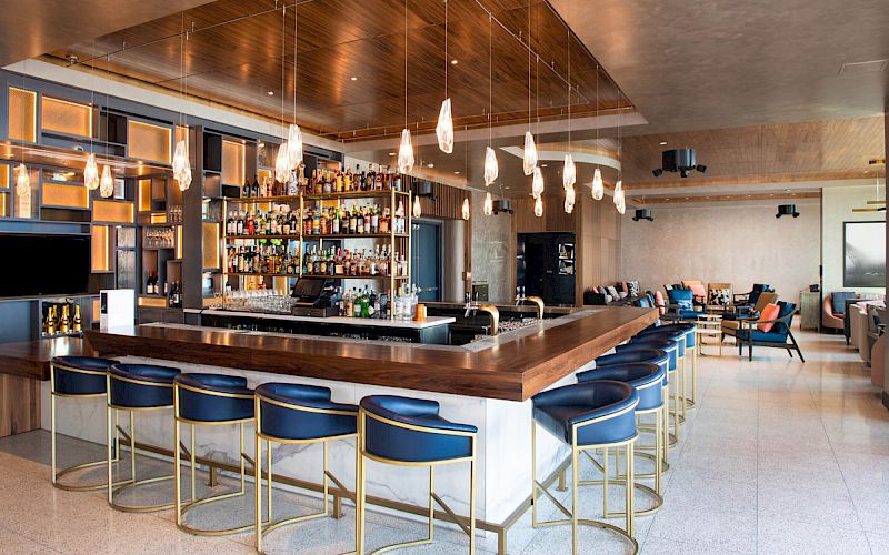 The image shows a modern bar with a wooden counter, blue stools, and various hanging lights, creating a stylish and inviting atmosphere.