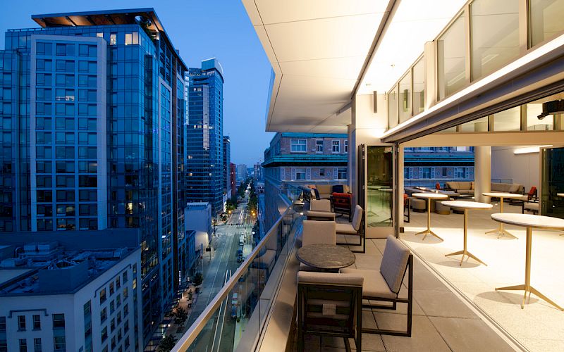 The image shows a modern rooftop terrace with seating overlooking a cityscape of tall buildings at dusk, creating an urban atmosphere.