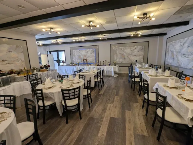 A bright banquet hall set with white-tablecloth tables and black chairs, lit by ceiling lights, with wall maps and marble artwork.