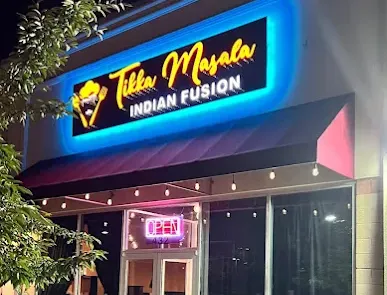 A night photo of a restaurant storefront reading &ldquo;Tikka Masala Indian Fusion&rdquo; with bright blue neon signage and glass doors.