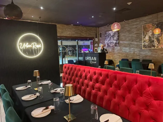 A cozy restaurant interior with a red tufted booth, black walls, neon &ldquo;Upper Room&rdquo; sign, and tables set with cutlery.