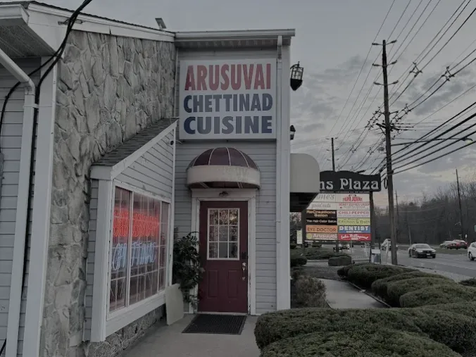 A shop storefront labeled &ldquo;Arusuvai Chettinad Cuisine&rdquo; with a red door and a nearby billboard by a street with power lines.